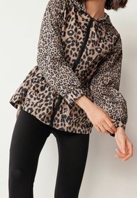 Personne portant une veste zippée imprimé léopard avec poignets élastiques et un pantalon noir, ajustant la manche sur un fond clair uni.