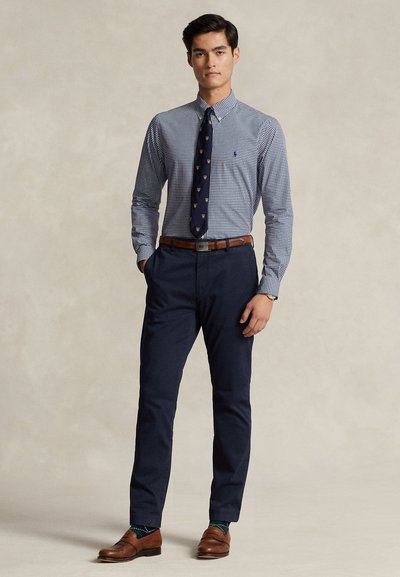 Junger Mann trägt blau kariertes Hemd, marineblaue Krawatte mit Muster, dunkelblaue Hose, braunen Gürtel und braune Loafers, steht mit der Hand in der Tasche.