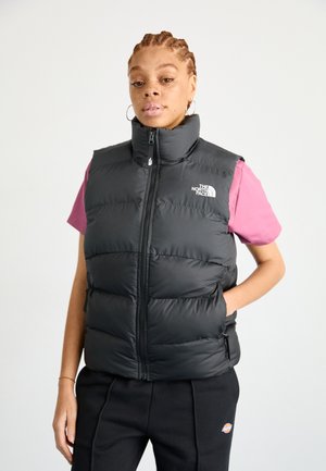 Schwarze Pufferweste mit hohem Kragen und Frontreißverschluss; verfügt über seitliche Reißverschlusstaschen und ein Logo auf der linken Brust. Tragen über einem pinken Shirt.