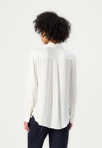 Langärmlige Bluse in Creme mit glatter Textur, Knopfleiste hinten und abgerundetem Saum. Mit Kragen und geknöpften Ärmeln.