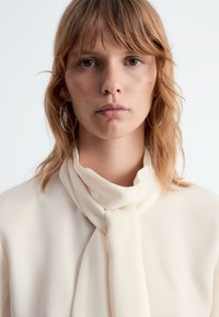 Cremfarbene Bluse mit einer sanften Textur, die mit einem gebundenen Halstuch und einem glatten, fließendenDesign versehen ist. Das Model hat helles, welliges Haar.