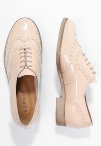 Chaussures oxford en cuir verni beige avec des détails perforés, bout rond, talon plat et fermeture à lacets, affichant une texture lisse et un design classique.
