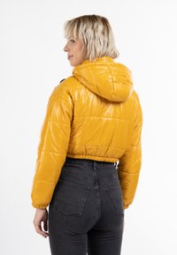 Gul puffjacka med en kortad design och huva, med en glänsande yta och horisontellt kviltat mönster, kombinerad med högmidjade svarta jeans.