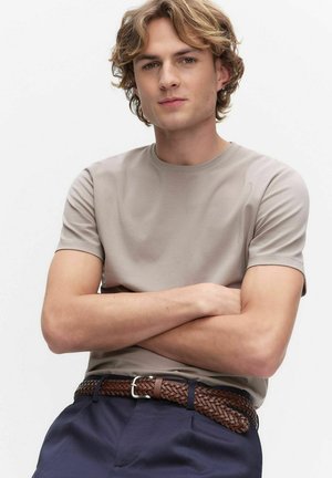 Ung mand med bølget blondt hår iført en beige kortærmet skjorte, marineblå bukser og et flettet brunt læderbælte, armene krydsede.