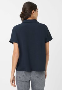 Polo shirt blu navy a maniche corte con vestibilità rilassata, caratterizzato da un colletto classico e una texture liscia, abbinato a jeans grigi con tasche posteriori.