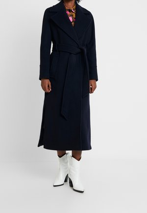 Person wearing a long dark navy belted coat, floral shirt, and white heeled ankle boots, standing against a plain background.