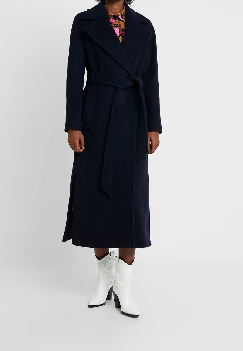 Personne portant un long manteau bleu marine foncé ceinturé, une chemise fleurie, et des bottines blanches à talons, se tenant devant un fond uni.