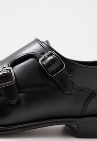 Scarpa in pelle nera con un design a doppia fibbia, texture liscia, tacco basso e cuciture rinforzate per una maggiore durabilità.