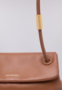Borsa a mano in pelle marrone con un design strutturato, dotata di un manico curvo e di un accento in hardware dorato. Texture liscia e logo leggermente impresso.