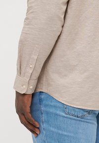 Camicia beige a maniche lunghe con polsini abbottonati, realizzata in un tessuto testurizzato. Abbinata a jeans in denim blu. La camicia ha un orlo arrotondato.