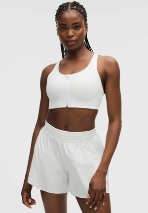 Frau mit geflochtenem Haar, die einen weißen Sport-BH mit Reißverschluss vorne und weiße Sportshorts trägt, steht vor einem schlichten hellen Hintergrund.