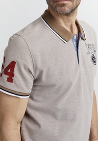 Polo beige con cuello marrón, detalles en negro y blanco, número "24" rojo en la manga y logo bordado en el pecho. Manga corta.