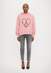 Svetlo roza flis hooded pulover z črnim potiskom srca in simbola miru, v kombinaciji s sivimi skinny jeanami in črnimi visokimi petami.