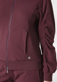 Bordeauxfärgad zip-up sweatshirt med ribbstickad nederkant och ärmslut, med detaljerade veck på ärmarna och en logotypaccent på den nedre sidan.