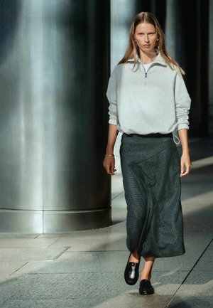 Woman walking indoors near large metal columns wearing a light gray quarter-zip pullover, dark gray long skirt, and black shoes.