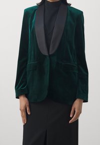 Smaragdgrön sammetsblazer med svart satin sjalkrage, två framfickor och långa ärmar, presenterad med en elegant, skräddarsydd silhuett.