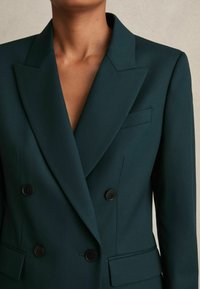 Zilā dubultā jaka ar asiem atloka galiem, smalku tekstūru un melnām pogām. Iespējams, ir viena krūšu kabata un strukturēts dizains.