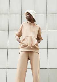 Tan spalvos Nike džemperis su raišteliais ir priekyje kišene, derinamas su atitinkamomis treniruočių kelnėmis. Baltos spalvos logotipo akcentai abiejuose. Minkšta, atsipalaidavusi siluetas.