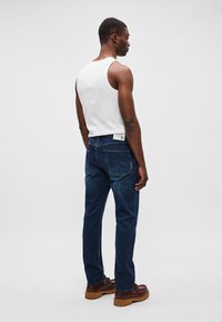 Jeans de culoare albastru închis, cu un croi drept, detalii cusute și două buzunare la spate. Purtați cu un maiou alb și pantofi maro.
