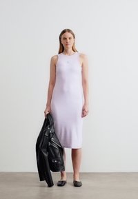 Γυναίκα με αμάνικο, μίντι φόρεμα σε ανοιχτό μοβ χρώμα, κρατώντας ένα μαύρο δερμάτινο μπουφάν στο δεξί της χέρι, στέκεται σε απλό πάτωμα μπροστά σε λευκό τοίχο.