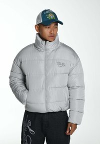 Grijze pufferjack met een hoge kraag, zigzagstiksels en een logo op de borst. Gepaard met donkere broeken met witte decoratieve tekst.