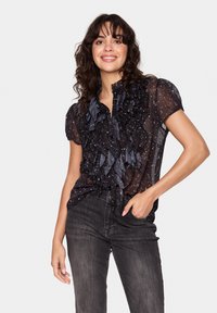 Črna prozorna bluzo z nabrano detajli, kratkimi rokavi in gumbnim zapiranjem spredaj. Ima zvezdasti vzorec in visok, nabran ovratnik.