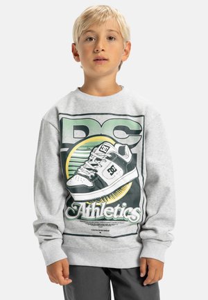 Blond kind in een grijze sweatshirt met een grote zwart-witte skateboard schoenafbeelding en de tekst "DC Athletics" aan de voorkant.