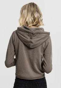 Brun cropped hoodie med en myk tekstur, med en stor hette og sømdetaljer. Bærer svarte jeans, viser bakvinkel.