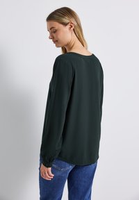 Mörkgrön blus med lång ärm, rund halsringning och lös passform. Tyget verkar slätt; har knäppta ärmslut och en böjd nederkant.