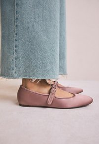 Ballerine in satin rosa con punta arrotondata e una cinturino di tessuto con una fibbia metallica circolare. Indossate con jeans azzurri strappati.