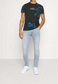 Svart t-shirt med batikmönster och blå detaljer, prydd med en tryckt logotyp, kombinerad med ljusblå jeans och vita sneakers. Avslappnad passform och korta ärmar.