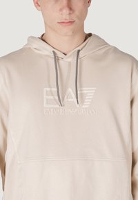 Felpa beige con cappuccio e coulisse elastica, caratterizzata da un logo testurizzato "EA7 EMPORIO ARMANI" sul petto, realizzata in un tessuto morbido e liscio.