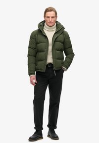 Giacca a vento verde con cappuccio, caratterizzata da una texture trapuntata, un colletto alto e tasche laterali, abbinata a un dolcevita crema e pantaloni neri.