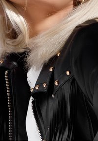 Chaqueta de cuero negra con tachuelas doradas y detalles de flecos, con un cuello de piel sintética. Textura suave y cierre de cremallera. Camisa blanca debajo.