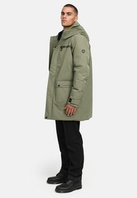 Olivnozeleni vodoodporni parka s kapuco, z sprednjim zadrgo, dvema velikima sprednjima žepoma in črnimi poudarki. Kombiniran s črnimi hlačami in čevlji.