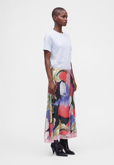 Femme aux cheveux courts portant un t-shirt blanc uni, une jupe midi fleurie colorée et des bottines noires à talons, debout de profil sur fond blanc.