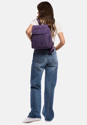 Zaino viola con superficie texturizzata, tasca frontale e spallacci regolabili. Indossato con jeans blu larghi e sneakers bianche.