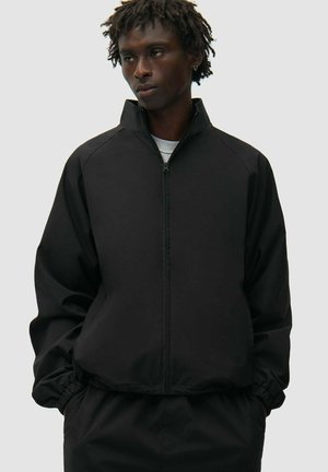 Giovane uomo dalla pelle scura e dreadlocks corte che indossa una giacca nera con zip e pantaloni neri abbinati, con le mani in tasca, guarda a sinistra.