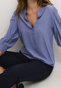 Blouse bleu clair avec un décolleté en V, des manches ballon et des poignets froncés. Le tissu a un motif texturé subtil. Portée avec un pantalon foncé.