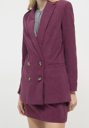 Woman wearing a double-breasted purple corduroy blazer with matching skirt and gray top against a white background.