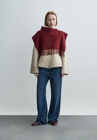 Béžový kabát z vlněné směsi s rozšířenými rukávy, kombinovaný s volnými modrými džínami. Červené pončo s třásněmi je přehodeno přes ramena.