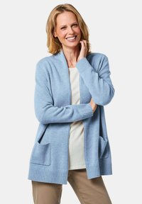 Hellblauer Cardigan, weicher Stoff, offene Vorderseite, gerippte Bündchen und Saum, zwei vordere Taschen, lockere Passform, über einem cremefarbenen Top getragen.