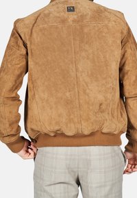 Ruggig bomberjacka i sandfärgad mocka med en mjuk textur, ribbade cuffs och nederkant, samt en märkesplåt vid kragen. Har en enkel, klassisk design.
