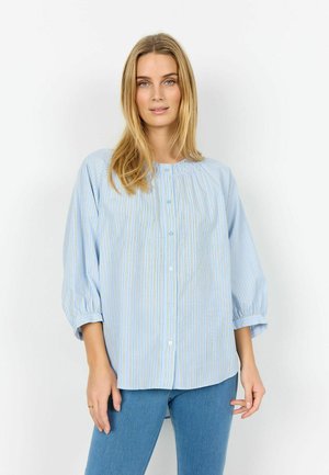 Blondynka ubrana w jasnoniebieską, długorękawową koszulę z cienkimi pionowymi paskami i niebieskie dżinsy, stojąca na białym tle.