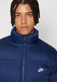 Μπλέι navy Nike μπουφάν με ψηλό γιακά, πλήρως μπροστινό φερμουάρ και λευκό λογότυπο στο αριστερό στήθος. Διαθέτει υφή με επενδεδυμένο σχέδιο.