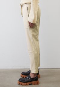 Beige tapered byxor med sidofickor med dragkedja och sömdetaljer, i kombination med kraftiga svarta skor med spänne och tjocka bruna sulor.