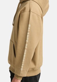 Beige hoodie made of cotton blend, featuring a kangaroo pocket, drawstring hood, and "BALANCE OF OPPOSITES" printed in white on the sleeve.