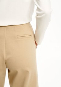 Pantaloni beige con una superficie liscia, dotati di una tasca posteriore slim con cuciture orizzontali. La parte superiore è una camicia bianca a maniche lunghe.