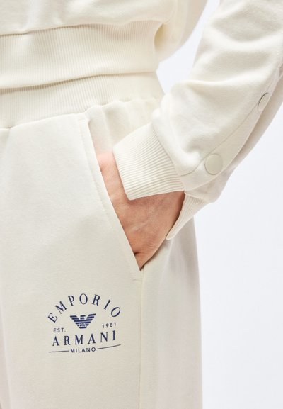 Χέρι στην πλαϊνή τσέπη κρεμ Emporio Armani φόρμας με μακρυμάνικο τοπ σετ που διαθέτει λεπτομέρειες με κουμπιά στο μανίκι.