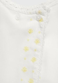 Vit plagg med gul blomsterbroderi och texturerad spetskant längs halsringningen, med klara knappar för stängning.
